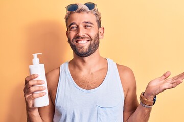 Young handsome blond man with beard on vacation holding bottle of sunscreen to protect skin celebrating achievement with happy smile and winner expression with raised hand