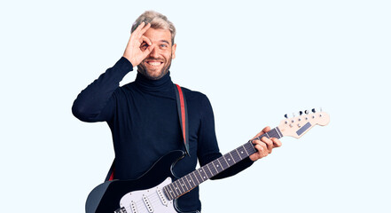 Young handsome blond man playing electric guitar smiling happy doing ok sign with hand on eye looking through fingers
