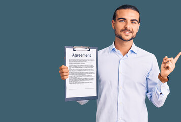 Young handsome man holding clipboard with agreement document smiling happy pointing with hand and finger to the side