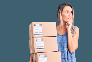 Young beautiful blonde woman holding delivery boxes serious face thinking about question with hand...