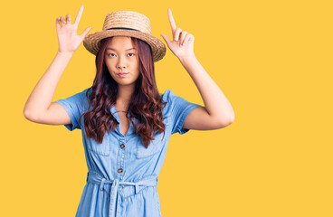 Obraz na płótnie Canvas Young beautiful chinese girl wearing summer hat doing funny gesture with finger over head as bull horns