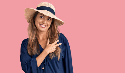 Middle age hispanic woman wearing summer hat cheerful with a smile of face pointing with hand and finger up to the side with happy and natural expression on face