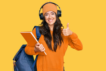 Beautiful brunette young woman wearing student backpack and holding books smiling happy and positive, thumb up doing excellent and approval sign