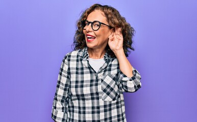 Middle age beautiful woman wearing casual shirt and glasses over isolated purple background smiling with hand over ear listening and hearing to rumor or gossip. Deafness concept.
