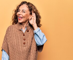 Middle age beautiful woman wearing casual sweater standing over isolated yellow background smiling with hand over ear listening and hearing to rumor or gossip. Deafness concept.