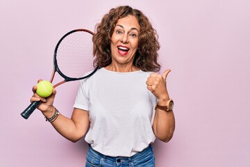 Middle age beautiful sportswoman playing tennis holding racket and ball over white background pointing thumb up to the side smiling happy with open mouth