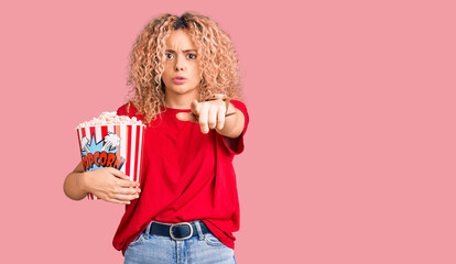 Young blonde woman with curly hair eating popcorn pointing with finger to the camera and to you, confident gesture looking serious