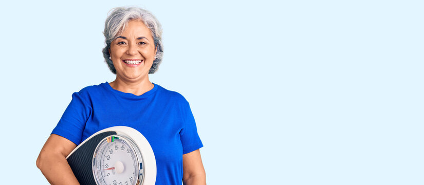 Senior woman with gray hair holding weight machine to balance weight loss looking positive and happy standing and smiling with a confident smile showing teeth