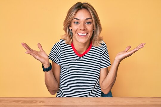 Beautiful caucasian woman wearing casual clothes sitting on the table celebrating victory with happy smile and winner expression with raised hands
