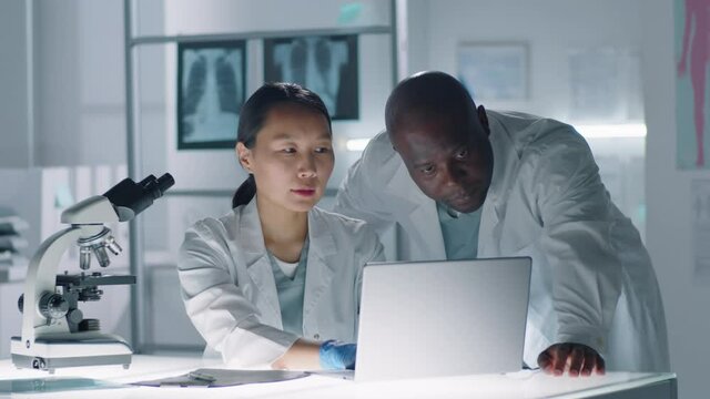 Two modern medical workers watching research data on laptop in laboratory then looking at camera