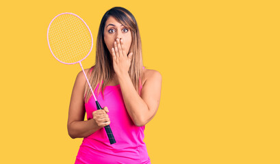 Young beautiful woman holding badminton racket covering mouth with hand, shocked and afraid for mistake. surprised expression