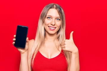 Naklejka premium Young beautiful blonde girl holding smartphone showing screen smiling happy and positive, thumb up doing excellent and approval sign