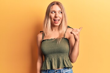 Young blonde woman wearing casual clothes pointing thumb up to the side smiling happy with open mouth