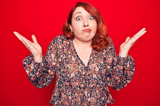 Young beautiful redhead woman wearing floral casual dress standing over red background clueless and confused with open arms, no idea and doubtful face.