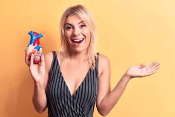 Young beautiful blonde woman asking for health care holding heart with veins and arteries celebrating achievement with happy smile and winner expression with raised hand