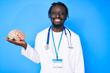 Young african american man with braids wearing doctor coat holding brain looking positive and happy...
