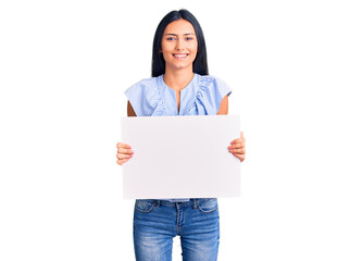 Young beautiful latin girl holding blank empty banner looking positive and happy standing and smiling with a confident smile showing teeth