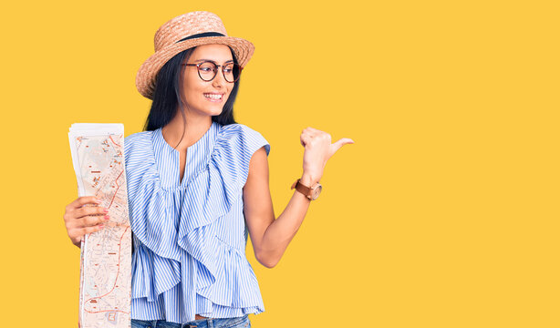 Young beautiful latin girl wearing summer hat holding map pointing thumb up to the side smiling happy with open mouth