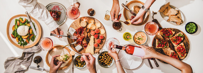 Friends wine and snacks party. Flay-lay of rose wine in glasses, cheese, fruit, meat, tomato brushettas, buratta salad and peoples hands over white table background, top view. Wine tasting concept - 377781004