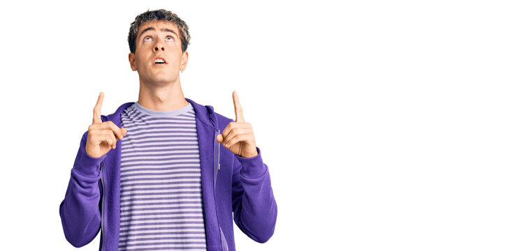 Young handsome man wearing casual purple sweatshirt amazed and surprised looking up and pointing with fingers and raised arms.