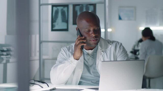 Modern African American lung specialist sitting at desk using laptop and talking on phone with his colleague