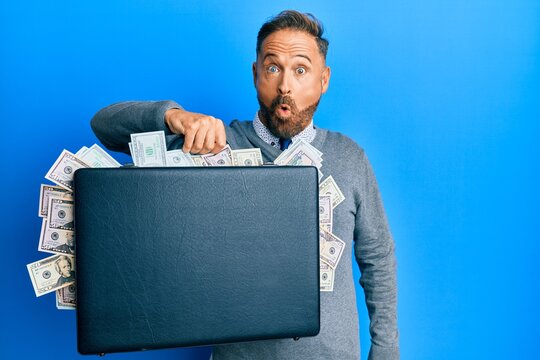Handsome middle age man holding briefcase full of dollars scared and amazed with open mouth for surprise, disbelief face