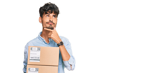 Young hispanic man holding delivery package serious face thinking about question with hand on chin,...