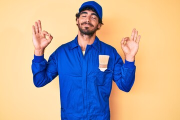 Naklejka premium Handsome young man with curly hair and bear wearing builder jumpsuit uniform relax and smiling with eyes closed doing meditation gesture with fingers. yoga concept.