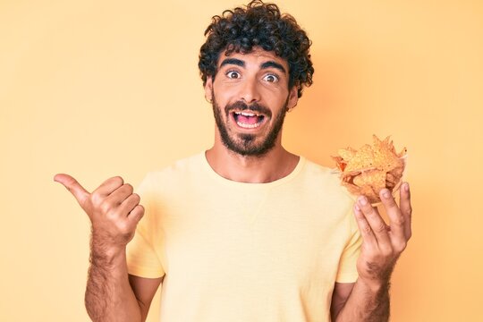 Handsome young man with curly hair and bear holding nachos potato chips pointing thumb up to the side smiling happy with open mouth