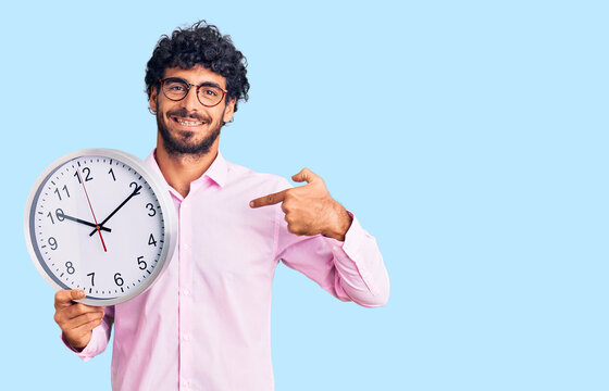 Handsome young man with curly hair and bear holding big clock pointing finger to one self smiling happy and proud