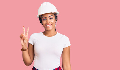 Young african american woman with braids wearing hardhat and builder clothes smiling with happy face winking at the camera doing victory sign. number two.