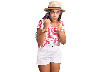 Obraz na płótnie Canvas Young african american woman with braids wearing summer hat ready to fight with fist defense gesture, angry and upset face, afraid of problem