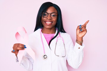 Young african american woman wearing doctor stethoscope holding pink cancer ribbon smiling happy pointing with hand and finger to the side