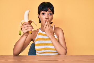 Brunette teenager girl eating banana as healthy snack covering mouth with hand, shocked and afraid for mistake. surprised expression