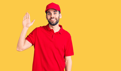 Young handsome man with beard wearing delivery uniform waiving saying hello happy and smiling, friendly welcome gesture