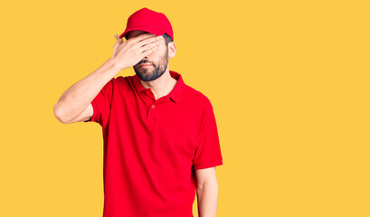 Young handsome man with beard wearing delivery uniform covering eyes with hand, looking serious and sad. sightless, hiding and rejection concept