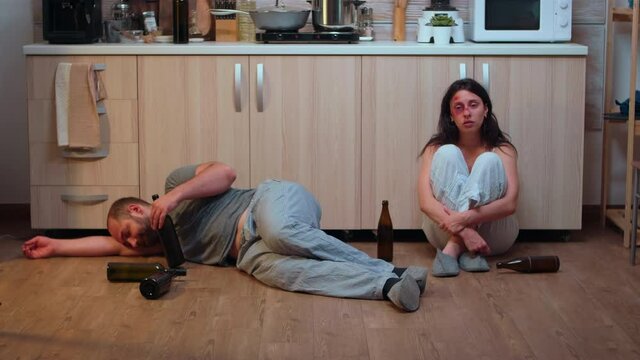 Alcoholic man lying on the floor near beaten wife surrounded by empty bottles. Drunk aggressive husband abusing injuring terrified helpless, afraid, beaten and panicked woman living in terror.