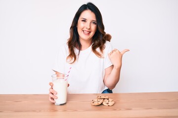 Young beautiful brunette woman drinking a glass of fresh milk with cookies pointing thumb up to the side smiling happy with open mouth