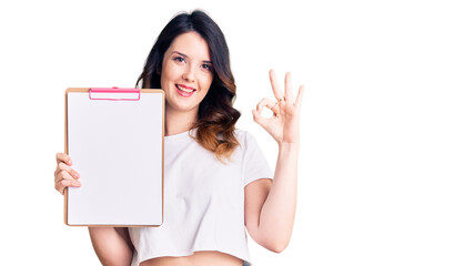 Fototapeta premium Beautiful young brunette woman holding clipboard with blank space doing ok sign with fingers, smiling friendly gesturing excellent symbol