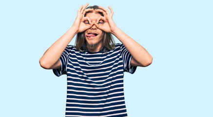 Handsome caucasian man with long hair wearing casual striped t-shirt doing ok gesture like binoculars sticking tongue out, eyes looking through fingers. crazy expression.