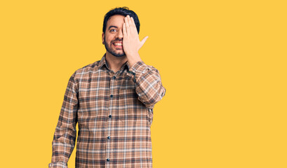Naklejka premium Young hispanic man wearing casual clothes covering one eye with hand, confident smile on face and surprise emotion.
