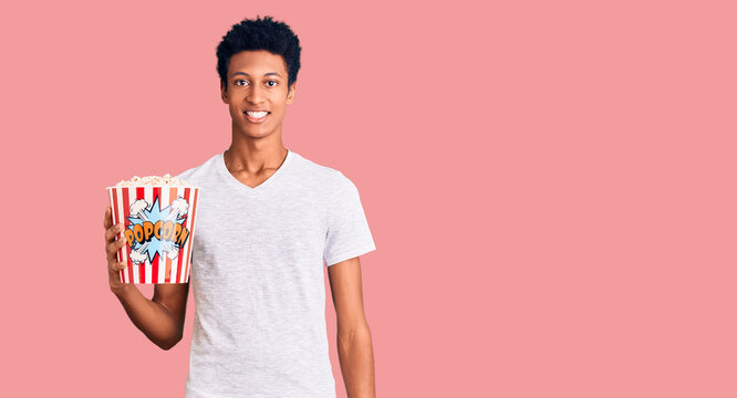 Young african american man holding popcorn looking positive and happy standing and smiling with a confident smile showing teeth