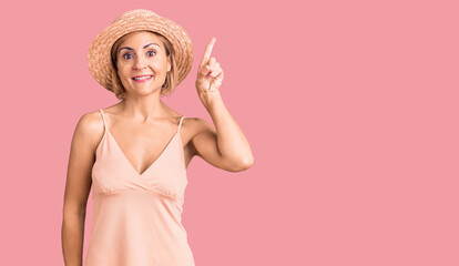 Young blonde woman wearing summer hat showing and pointing up with finger number one while smiling confident and happy.