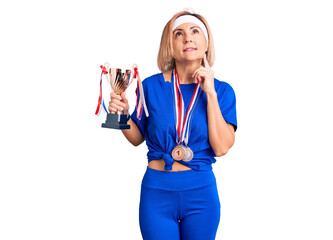 Young blonde woman holding champion trophy wearing medals serious face thinking about question with hand on chin, thoughtful about confusing idea