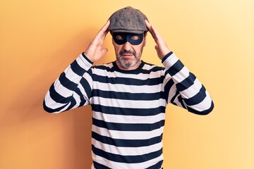 Middle age handsome burglar man wearing cap and mask over isolated yellow background with hand on head, headache because stress. Suffering migraine.