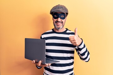 Middle age handsome burglar man wearing cap and mask using laptop over yellow background smiling happy and positive, thumb up doing excellent and approval sign