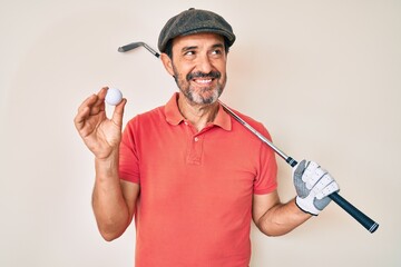 Middle age hispanic man holding golf club and ball smiling looking to the side and staring away thinking.