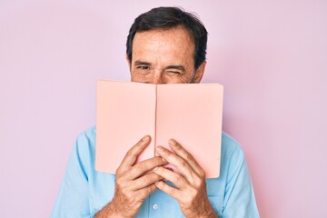 Middle age hispanic man reading book winking looking at the camera with sexy expression, cheerful...