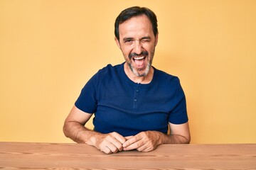 Middle age hispanic man wearing casual clothes sitting on the table winking looking at the camera...