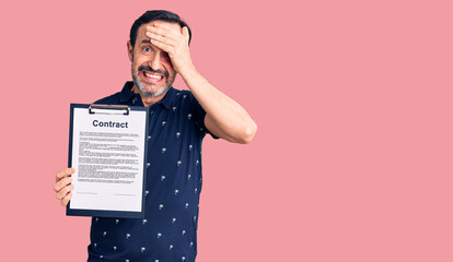 Middle age handsome man holding clipboard with contract document stressed and frustrated with hand on head, surprised and angry face